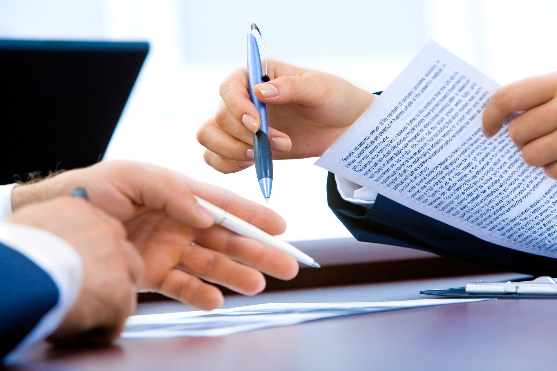 Un homme et une femme en costumes de travail dont on ne voie que les mains débattent. La femme tient en main un document imprimé et un stylo. Manifestement , elle explique ou argumente